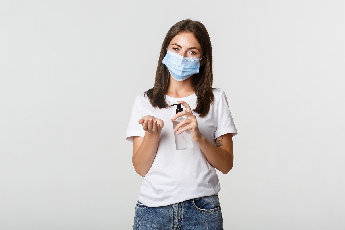 Covid-19, health and social distancing concept. Attractive young brunette woman in medical mask applying hand sanitizer on hand, white background